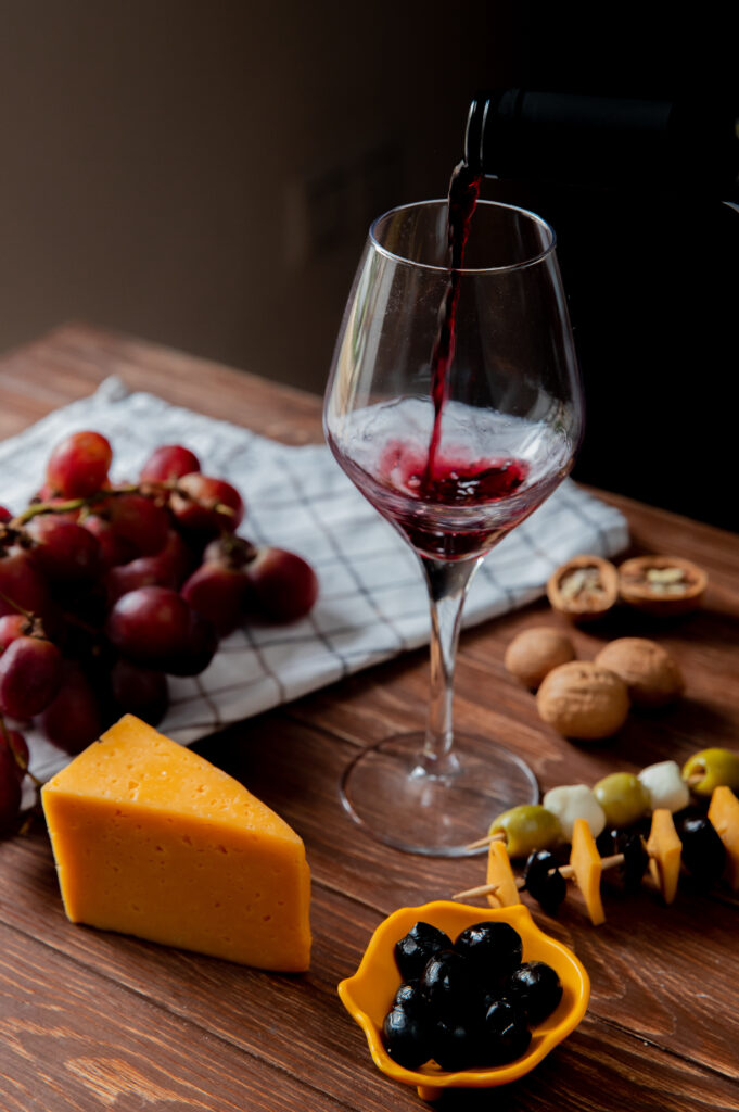 side view of red wine pouring into glass with cheddar and parmesan cheese olive walnut grape on wooden surface and black background